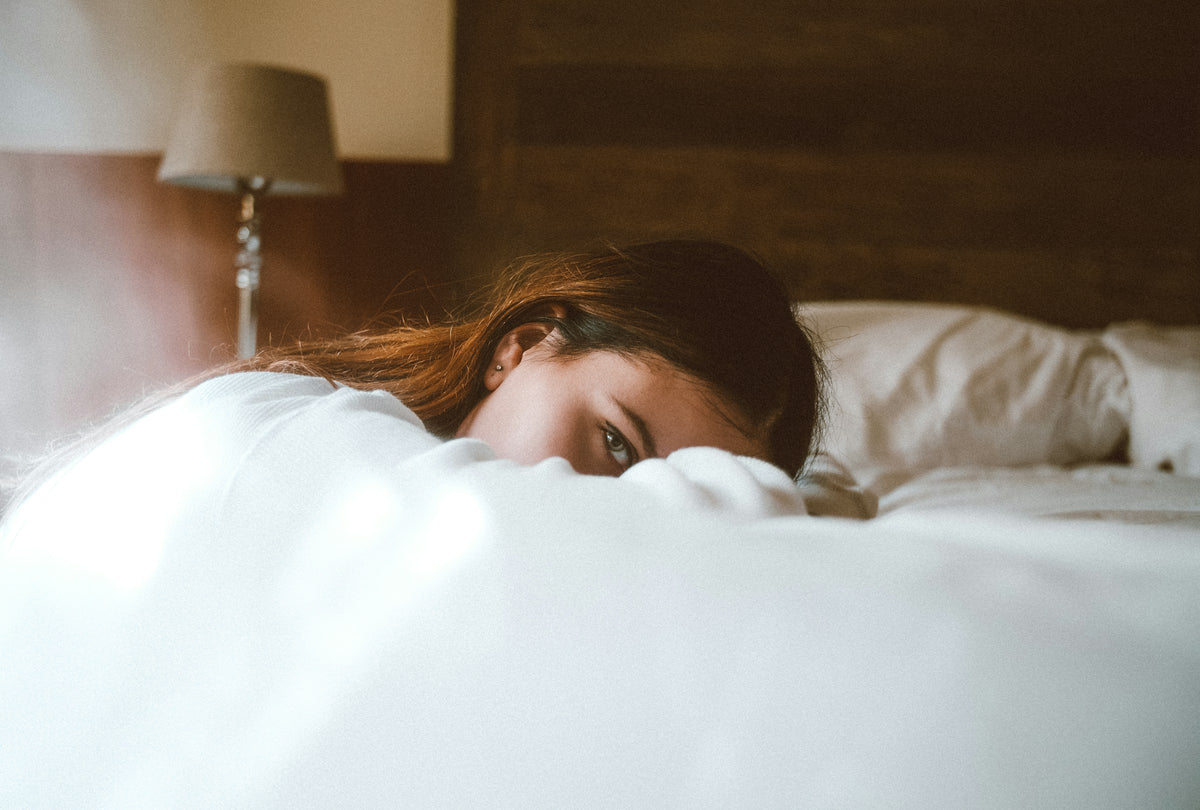 Femme allongée dans un lit aux draps blancs, sous la lumière tamisée d’une lampe de chevet